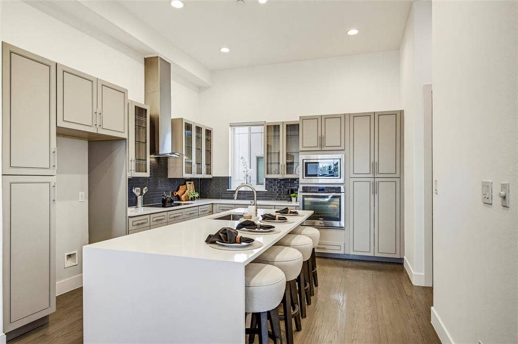 Kitchen featuring light-toned cabinetry with glass-front accents, an expansive quartz-topped island, stainless steel built-in double ovens, a chimney-style range hood, and wood-finish flooring