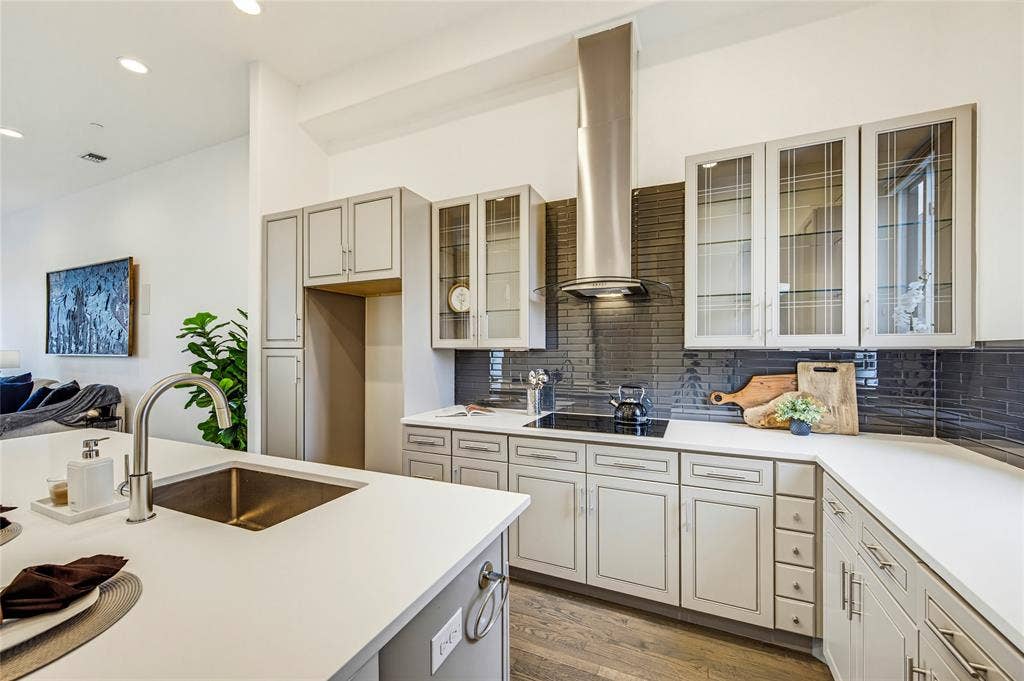Kitchen featuring custom cabinetry with glass-front uppers, solid-surface countertops, a stainless steel chimney-style range hood, high-gloss subway tile backsplash, and an integrated island sink