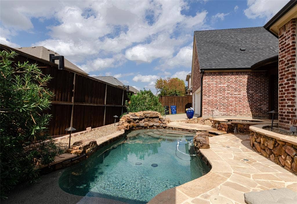 In-ground pool featuring natural stone coping, an integrated rock waterfall, and flagstone decking surrounded by a wood privacy fence and masonry exterior wall