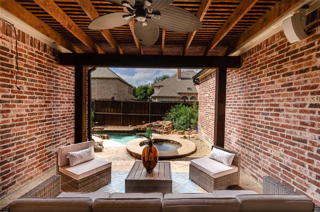 Covered patio featuring exposed wood-beam ceiling, ceiling fan, and exposed brick walls, overlooking a swimming pool and built-in stone spa