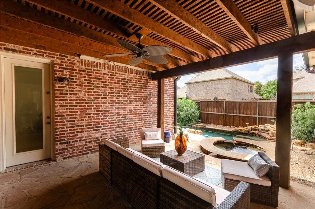 Covered outdoor patio featuring a slatted wood ceiling, exposed brick wall, stone-paved flooring, built-in ceiling fan, and access to a swimming pool with a stone-surround spa