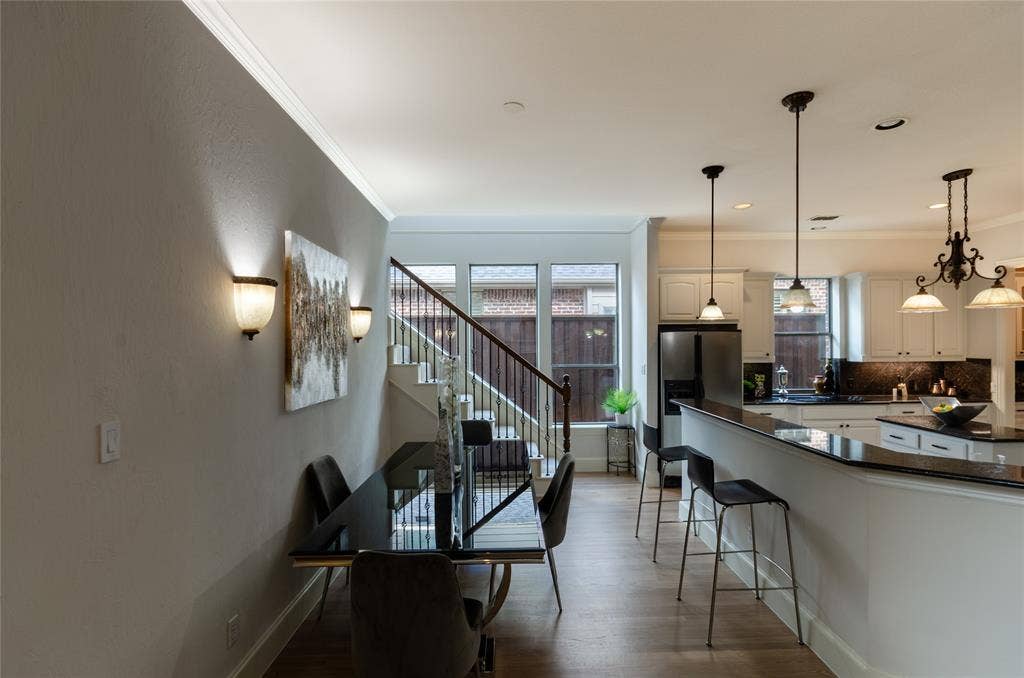 Open-concept interior featuring light wood-finish flooring, white cabinetry, polished dark stone countertops, decorative pendant lighting, and a staircase with wrought iron balusters