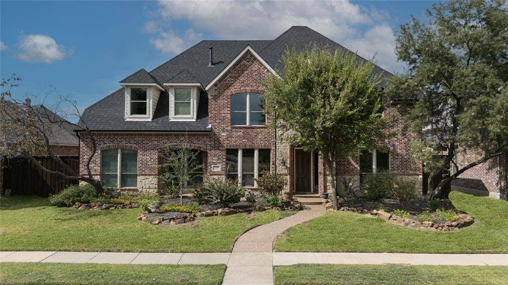Two-story exterior featuring multi-toned brick and stone masonry, a dark shingle roof, arched window detailing, dormers, and a paved walkway