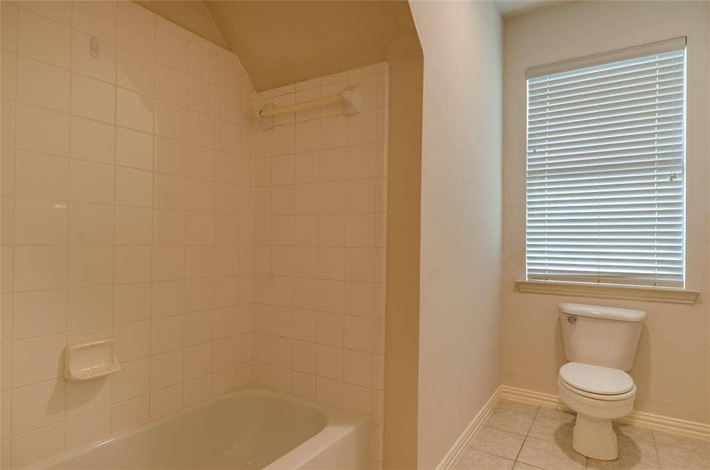 Bathroom featuring a porcelain tub with white tile surround, built-in soap dish, integrated grab bar, two-piece toilet, and window with horizontal blinds