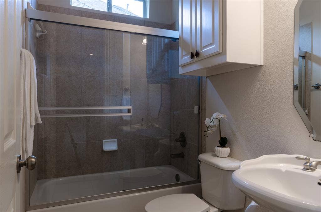 Full-height tiled shower surround, sliding glass enclosure, white upper wall cabinet, and pedestal sink with metallic fixtures