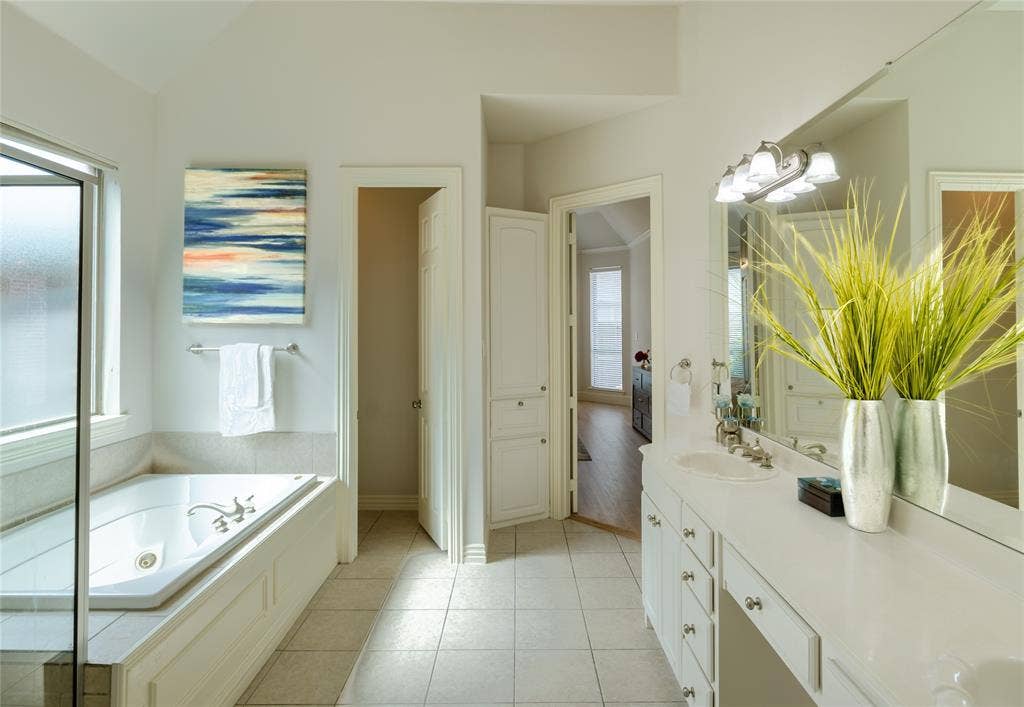 Bathroom featuring a deep soaking tub with a tile surround, built-in cabinetry, dual-sink vanity with white countertops, expansive wall-to-wall mirror, and neutral floor tile