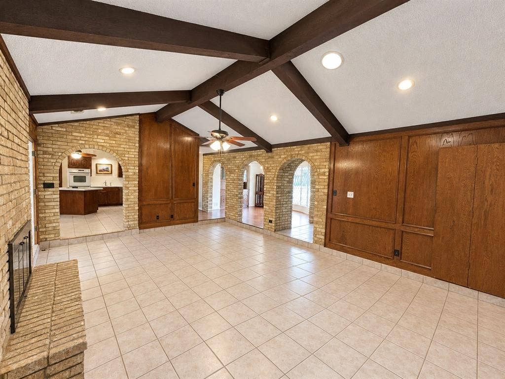 3408 Bellah Court, Irving, TX 75062 Vaulted ceiling featuring exposed dark-wood beams, brick-accented arched openings, full-wall wood paneling, tiled flooring, and a floor-to-ceiling masonry fireplace