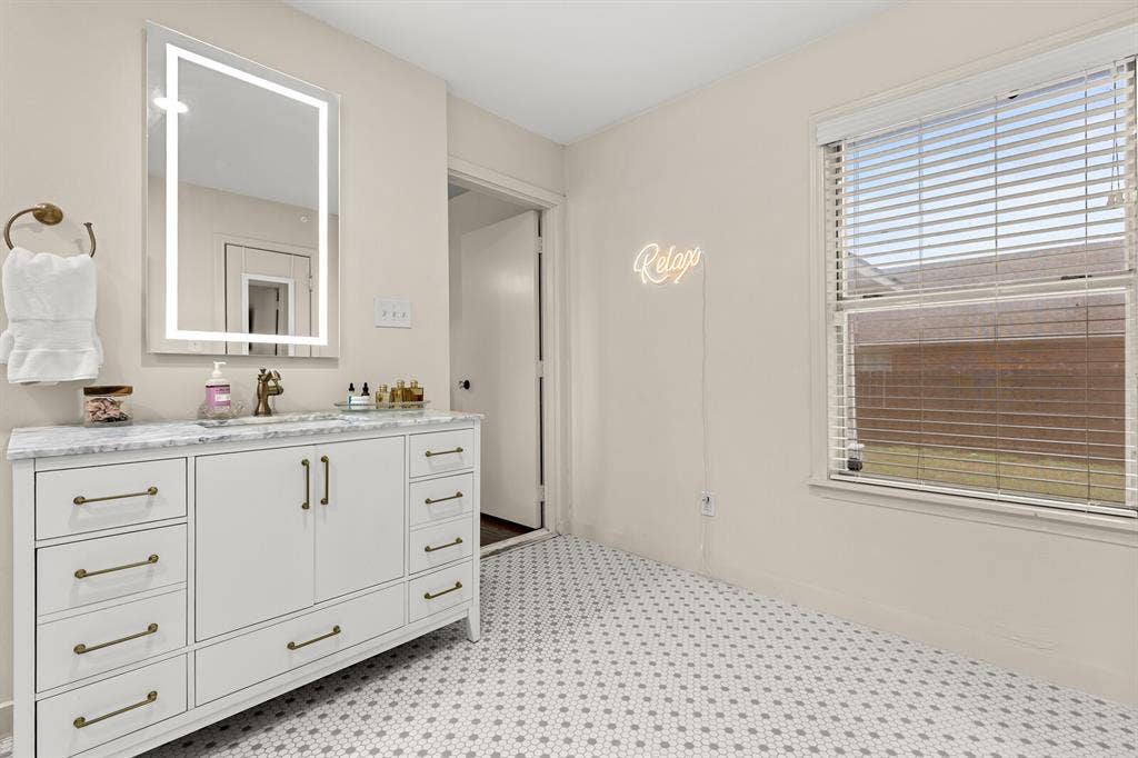2425 S 2nd Street, Waco, TX 76706 White vanity cabinet featuring brass hardware and a stone-composite countertop, illuminated by an integrated LED-frame mirror, complemented by penny-tile flooring and horizontal window blinds
