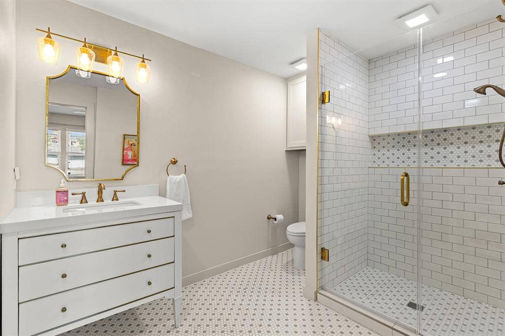 2425 S 2nd Street, Waco, TX 76706 Bathroom featuring a vanity with a white countertop, gold-toned hardware, wall-mounted four-bulb light fixture, glass-enclosed shower with subway tile surround, and patterned tile flooring