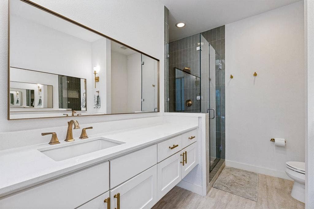 2107 Bennett Avenue, #20, Dallas, TX 75206 White shaker-style cabinetry with gold-tone hardware, crisp quartz countertop, expansive wall-mounted mirror, walk-in shower with vertical tile surround and glass enclosure, and wood-finish flooring