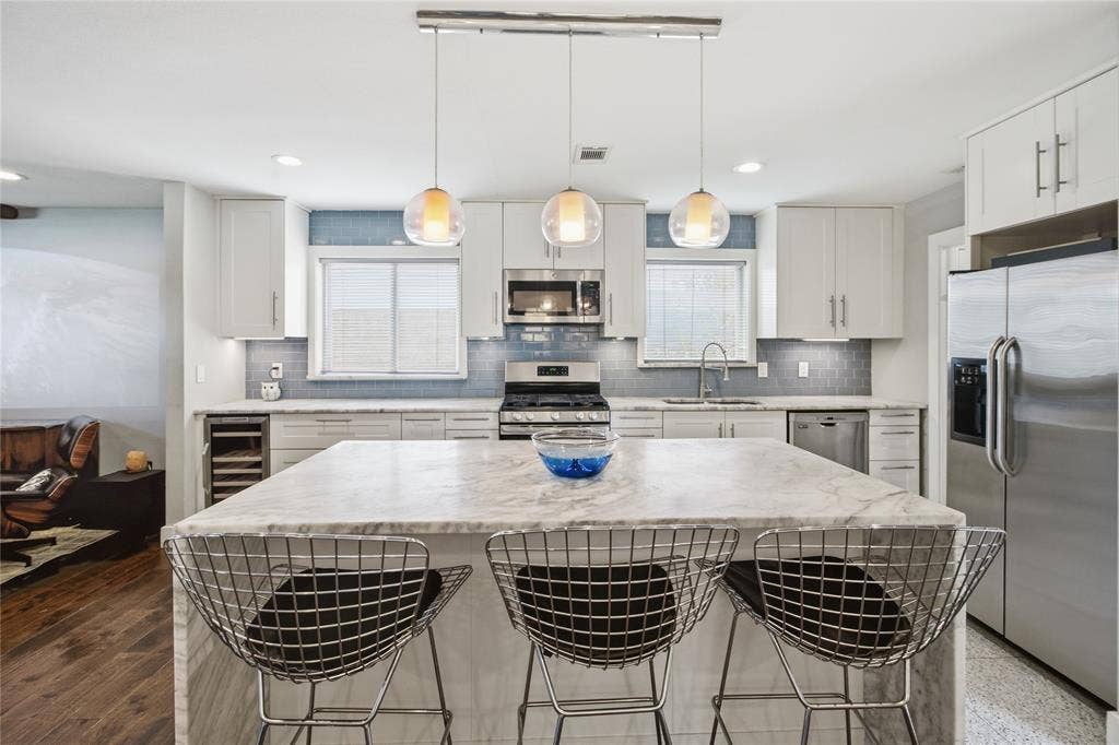 2712 Beechmont Place, Dallas, TX 75228 The beautifully updated kitchen features an expansive quartz waterfall island with seating for three, crisp white shaker-style cabinetry, stainless steel appliances, and a soft blue subway tile backsplash. Three glass pendant lights complete the contemporary design.