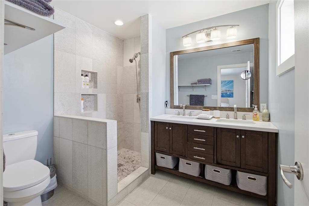 2712 Beechmont Place, Dallas, TX 75228 The primary ensuite bathroom features a spacious walk-in shower with tile surround, pebble-style flooring, and recessed storage niches. A double-sink vanity with dark wood cabinetry, generous counter space, and open shelving provides everyday convenience in a contemporary setting.