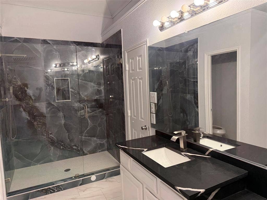 14 Heritage Way, Allen, TX 75002 Walk-in shower featuring large-format dark patterned tile and glass enclosure, vanity with dark stone countertop, undermount sink, and multi-bulb vanity lighting
