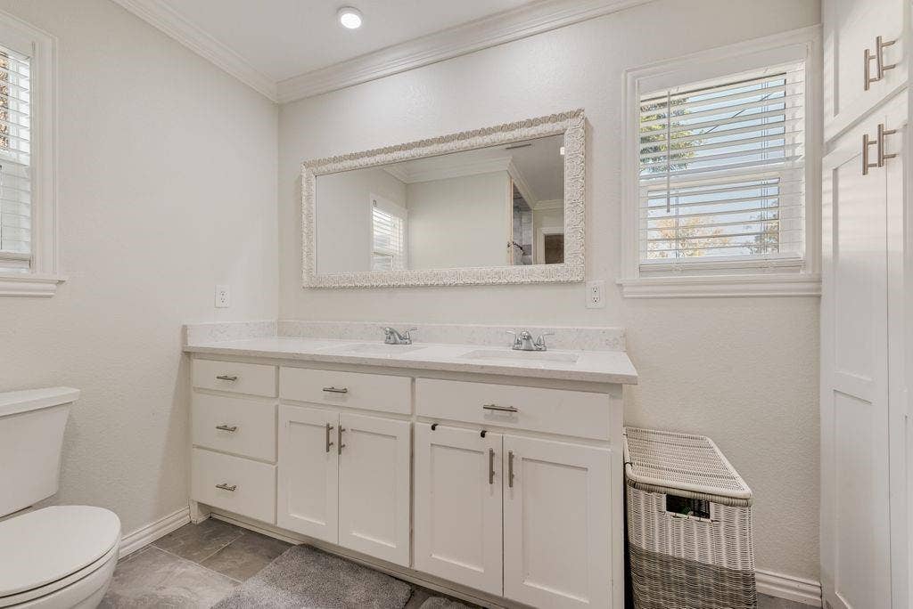 2815 Anzio Drive, Dallas, TX 75224 Dual-sink vanity with a stone-surface countertop, white shaker-style cabinetry, crown molding, framed wall mirror, and integrated floor-to-ceiling storage cabinetry