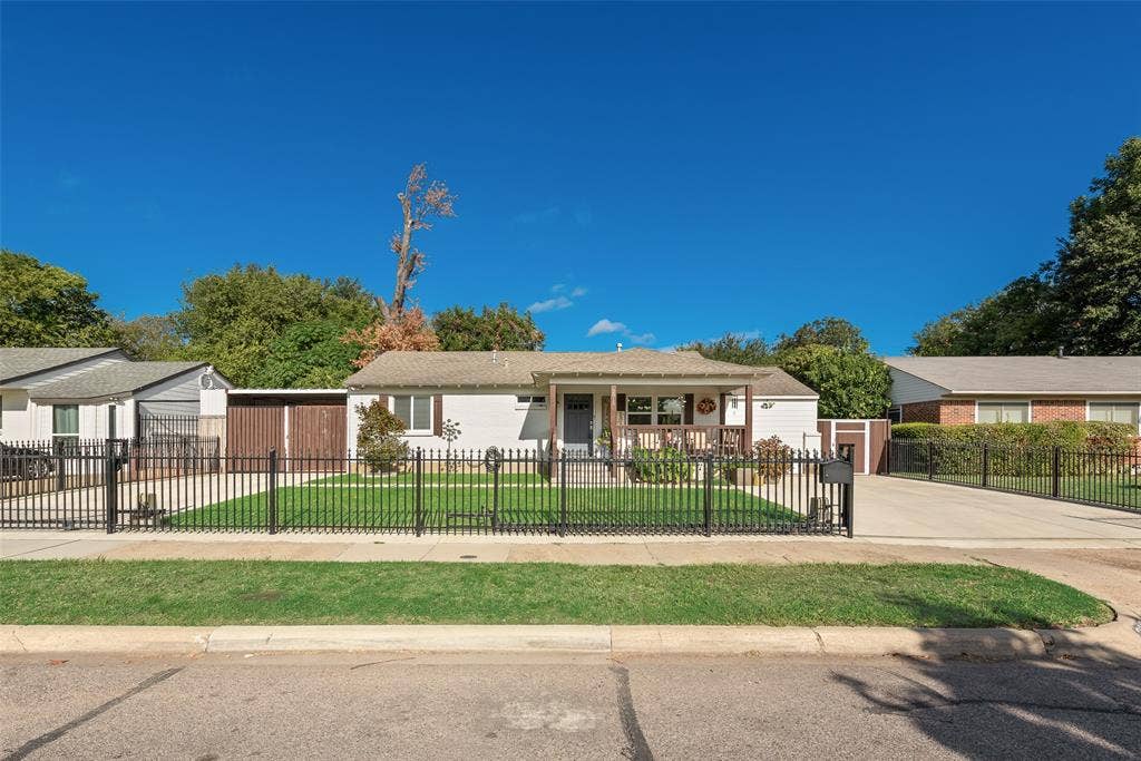2815 Anzio Drive, Dallas, TX 75224 Single-story residence featuring a light-colored exterior, covered front porch with a wood-finished railing, metal perimeter fencing, and an attached carport
