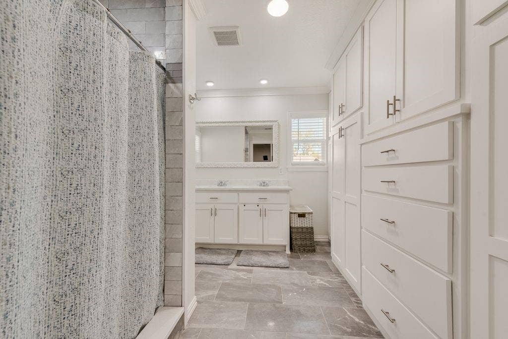 2815 Anzio Drive, Dallas, TX 75224 Bathroom featuring large-format tile flooring, extensive white built-in cabinetry with brushed nickel hardware, a dual-sink vanity, and a tub enclosure with subway-tiled walls