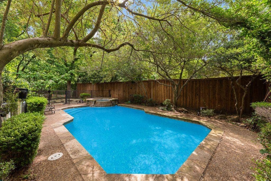 1418 Yakimo Drive, Dallas, TX 75208 In-ground swimming pool featuring a stone-accented spillover spa, flagstone decking, wood privacy fencing, and mature canopy trees