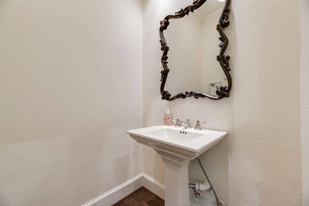 1418 Yakimo Drive, Dallas, TX 75208 White pedestal sink with a dual-handle metal faucet, complemented by an ornate, scrolled-edge wall mirror and dark wood-finish flooring