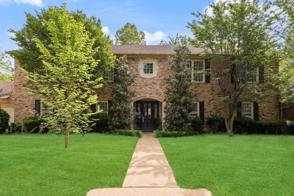 1418 Yakimo Drive, Dallas, TX 75208 Two-story brick exterior featuring a stone arched entryway, symmetrical multi-pane windows with dark shutters, a decorative central oval window, and a paved walkway