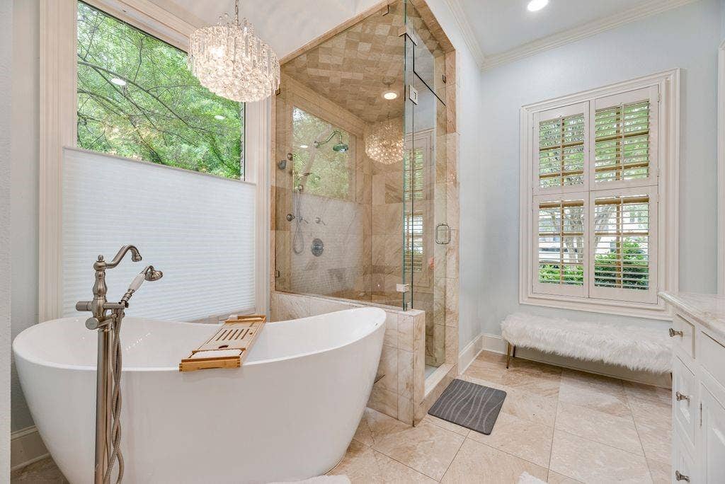 1418 Yakimo Drive, Dallas, TX 75208 Freestanding soaking tub with floor-mounted brushed nickel faucet, glass-enclosed shower with stone tile surround, multi-pane windows with integrated shutters, and crystal chandelier fixture