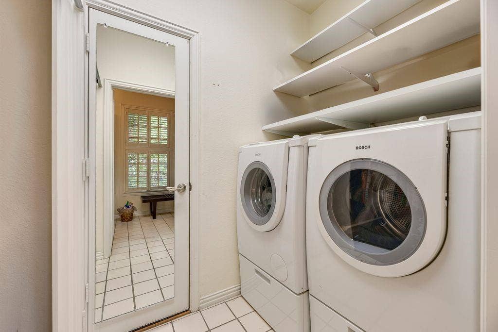 1418 Yakimo Drive, Dallas, TX 75208 Laundry room featuring Bosch white front-load washer and dryer, white ceramic tile flooring, and overhead built-in shelving