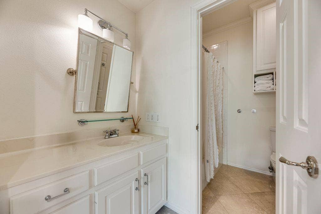 1418 Yakimo Drive, Dallas, TX 75208 White vanity with an integrated countertop, polished chrome hardware, wall-mounted mirror, three-light fixture, and built-in shelving unit