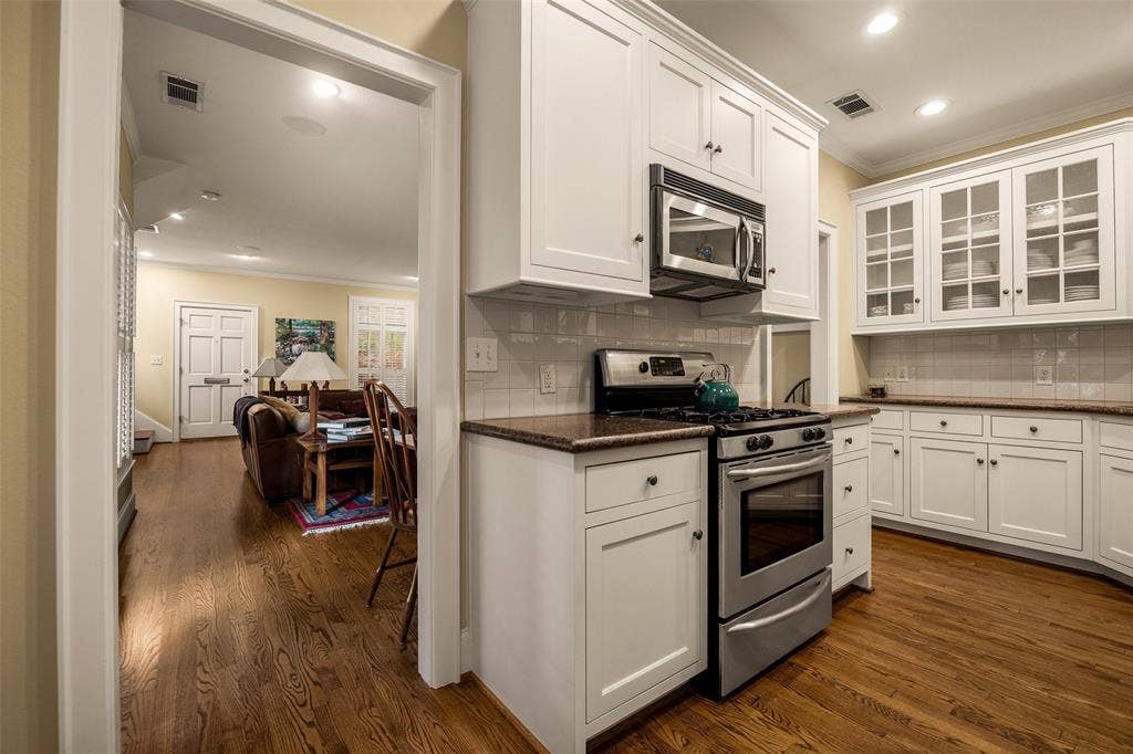 6020 Tremont Street, Dallas, TX 75214 Kitchen featuring white shaker-style cabinetry, stainless steel appliances, glass-front upper cabinets, stone-surfaced countertops, and wood-finish flooring