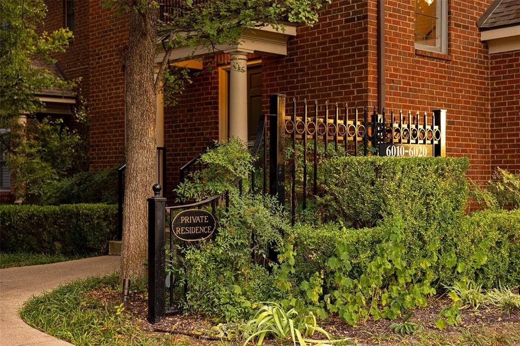 6020 Tremont Street, Dallas, TX 75214 Exterior facade featuring red brick masonry, decorative metal fencing, a covered entryway with column support, and landscaped garden beds