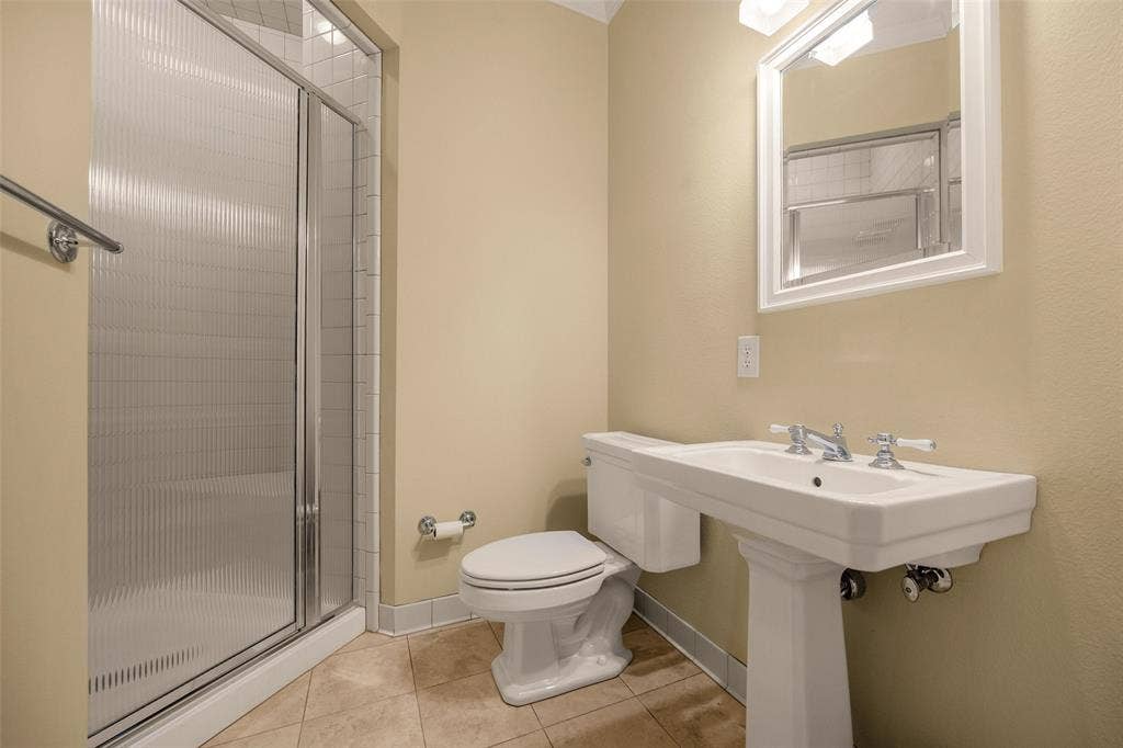 6020 Tremont Street, Dallas, TX 75214 Shower with semi-frameless fluted glass doors and tiled surround, pedestal sink with cross-handle faucet, white framed wall mirror, and neutral-tone tile flooring