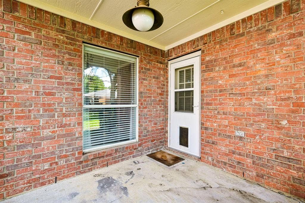 758 Livingston Drive, Allen, TX 75002 Covered patio featuring red brick masonry walls, a single-lite glass entry door with an integrated pet portal, a large window with white-trimmed casing, and a dome-style overhead pendant light