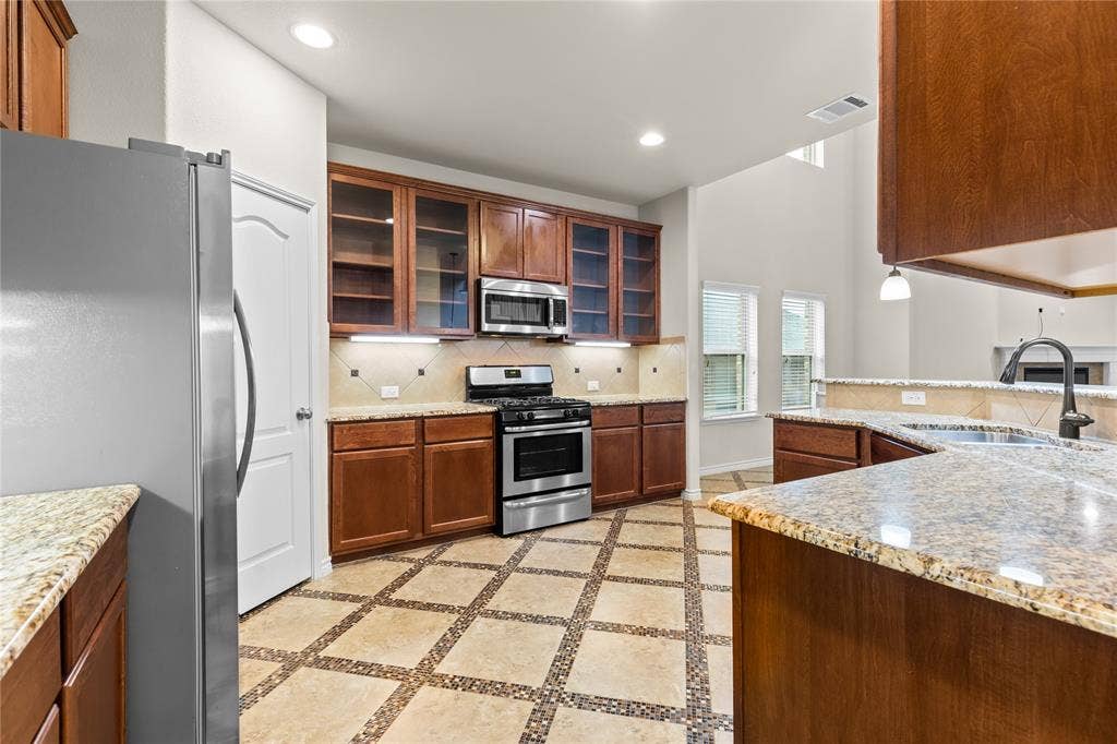 5751 Mountain Hollow Drive, Dallas, TX 75249 Kitchen featuring warm-toned wood cabinetry with glass-front uppers, stone-patterned countertops, stainless steel appliances, and tile flooring with decorative mosaic inlay