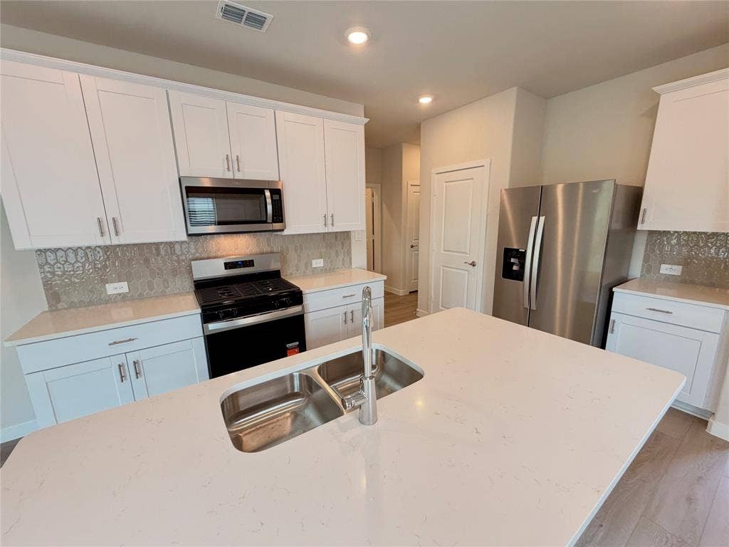 Kitchen featuring white cabinetry, quartz countertops, a central island with an undermount dual-basin sink, patterned tile backsplash, and stainless steel appliances