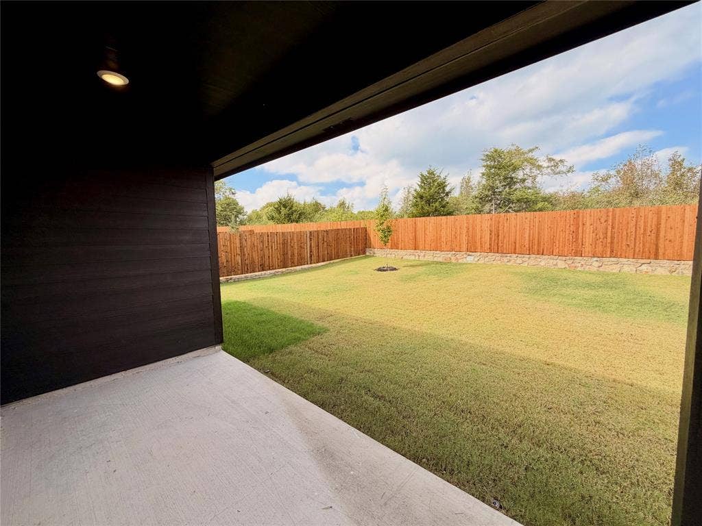 Covered patio with recessed lighting, concrete flooring, and dark-toned exterior siding, opening to a fenced backyard with a stone retaining wall