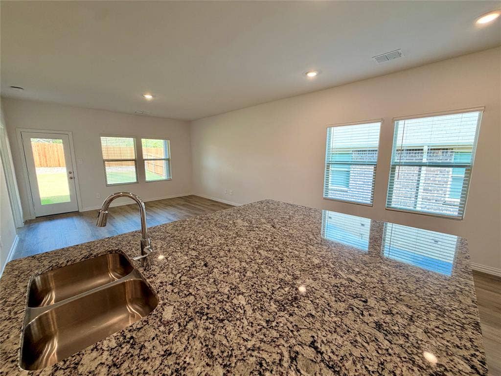Kitchen island featuring a stone countertop, dual-basin stainless steel sink, gooseneck faucet, wood-finish flooring, and natural light via multiple windows