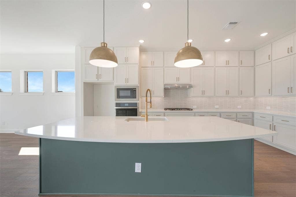 3469 Jersey Drive, Midlothian, TX 76065 Kitchen island with stone-surface countertop, white cabinetry with silver-tone hardware, built-in microwave and wall oven, brass-finish faucet, and pendant lighting