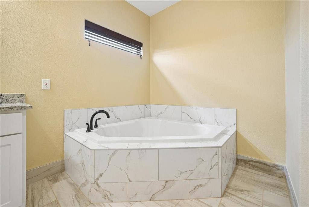 9060 Mitchell Bend Court, Granbury, TX 76048 Corner soaking tub featuring a stone-look tile surround, white cabinetry, a stone countertop, and a matte black faucet