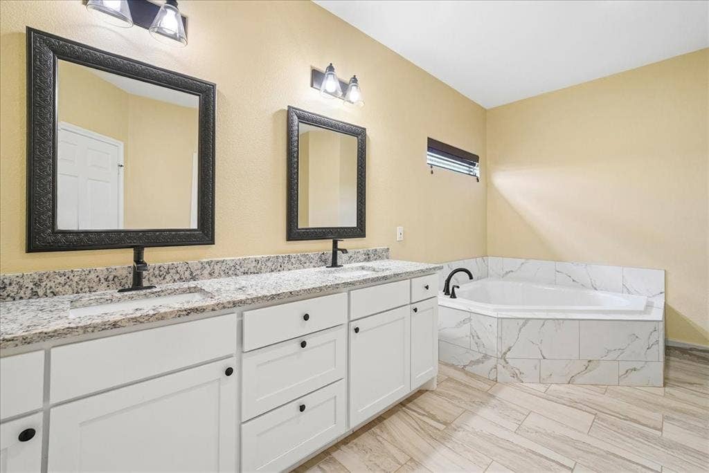 9060 Mitchell Bend Court, Granbury, TX 76048 Dual vanity with light-toned stone-finish countertop, white shaker-style cabinetry, framed wall mirrors, corner soaking tub with surround tile, and tile flooring