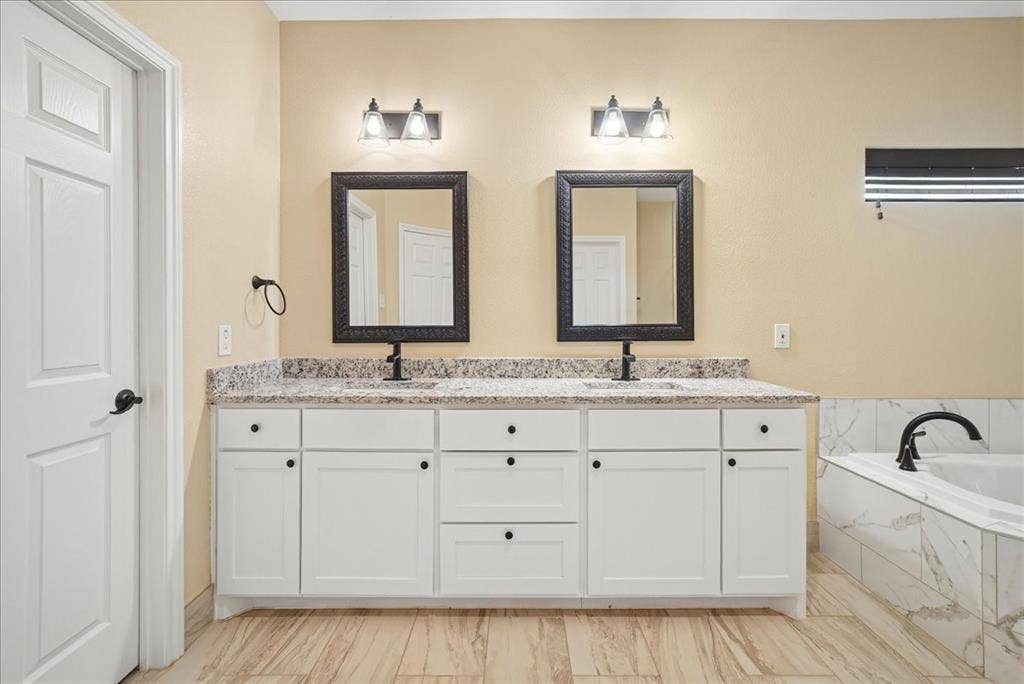 9060 Mitchell Bend Court, Granbury, TX 76048 Double vanity with white shaker-style cabinetry, stone-finish countertop, dual under-mount sinks, dark-framed mirrors, and wood-finish flooring