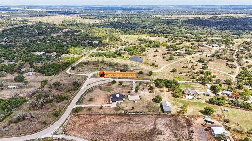 9060 Mitchell Bend Court, Granbury, TX 76048 Aerial perspective of a sprawling property featuring expansive acreage, a metal-roofed residence, a detached secondary structure, and established tree clusters
