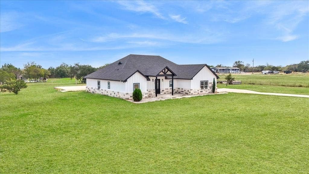 9060 Mitchell Bend Court, Granbury, TX 76048 Single-story exterior featuring white vertical siding, stone masonry wainscoting, a dark shingle roof, and a covered front entry with timber-style accents, situated on expansive grounds