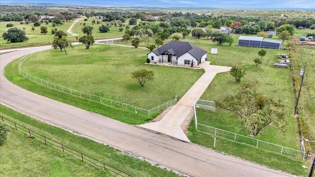 9060 Mitchell Bend Court, Granbury, TX 76048 Single-story residence featuring a light-toned exterior, dark shingle roof, concrete driveway, perimeter metal fencing, and an auxiliary detached structure on a sprawling acreage lot