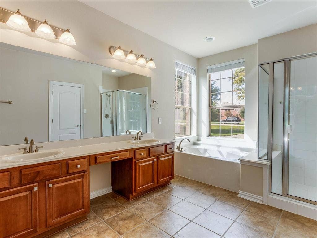 Dual-sink vanity with wood-finish cabinetry, solid-surface countertops, soaking tub, glass-enclosed shower, and tile flooring