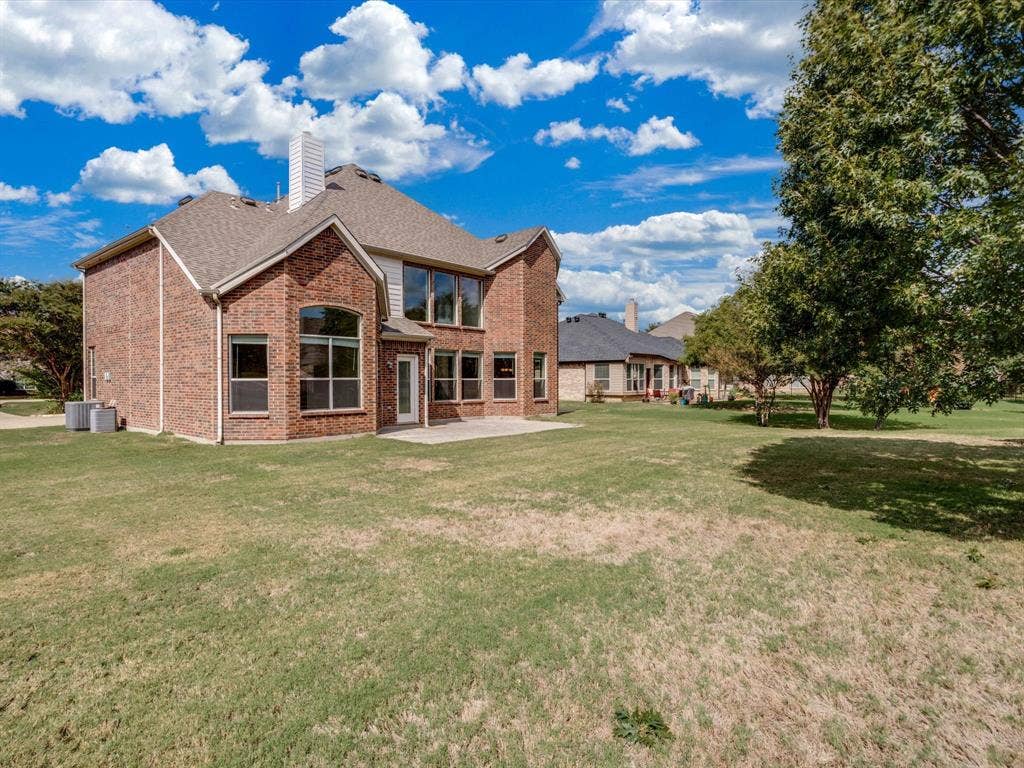 Two-story brick exterior featuring a multi-gabled roofline, expansive lawn, concrete patio, and large glass-paneled windows