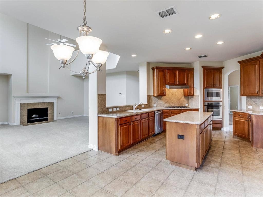 Kitchen featuring wood cabinetry, stone-finish countertops, tile backsplash, built-in wall oven, and a center island, complemented by a fireplace with stone surround