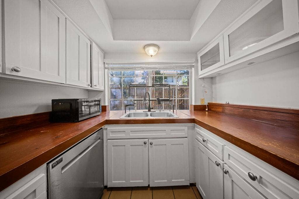 738 Intrepid Drive, Garland, TX 75043 Kitchen featuring white shaker-style cabinetry, wood-finish countertops, stainless steel dual-basin sink, upper glass-front cabinets, and a window with a grid pattern