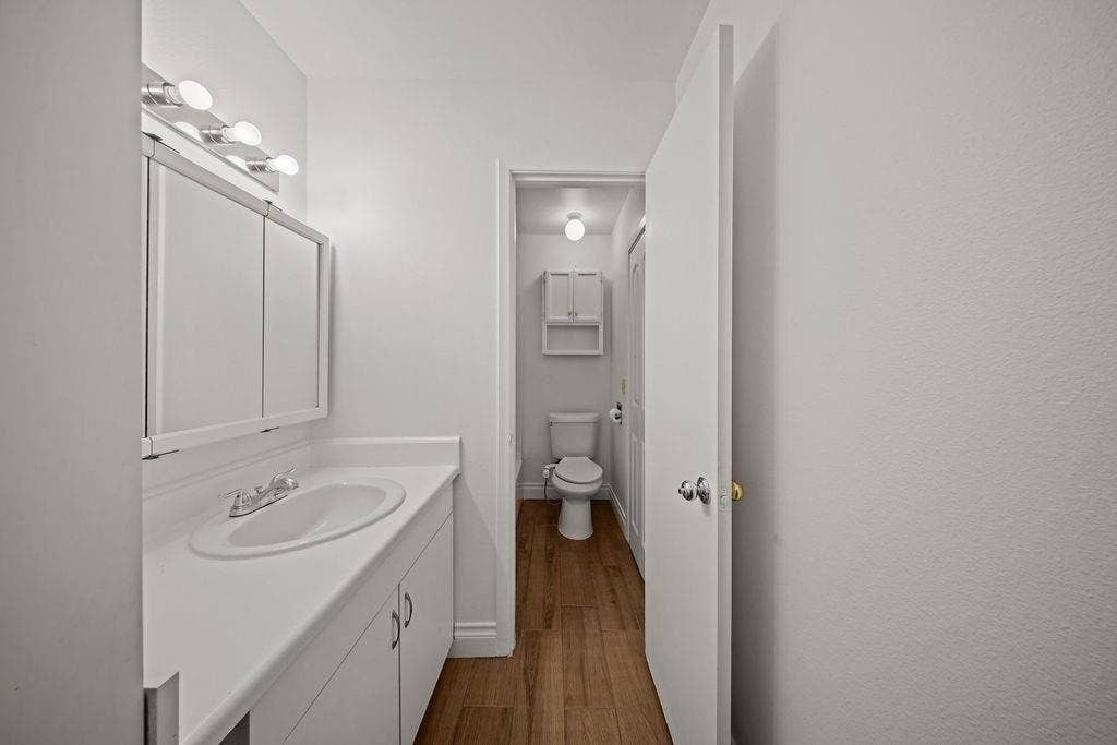 738 Intrepid Drive, Garland, TX 75043 Bathroom featuring a white vanity cabinet with an integrated sink, a mirrored medicine cabinet, a multi-bulb light fixture, wood-finish flooring, and a wall-mounted storage cabinet above the toilet