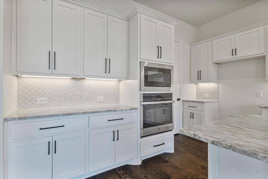 2817 Yarmouth Street, Celina, TX 75009 Kitchen featuring white Shaker-style cabinetry, herringbone-patterned tile backsplash, light-toned stone countertops, built-in stainless steel double ovens, and dark wood-finish flooring