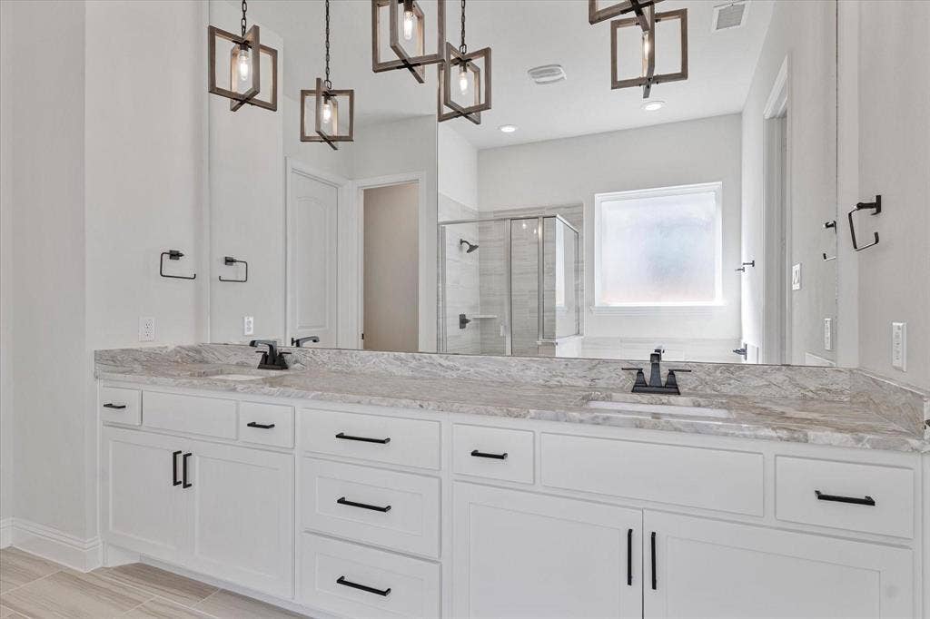 2817 Yarmouth Street, Celina, TX 75009 Dual-sink vanity with stone-finish countertop, white Shaker-style cabinetry, matte black hardware, geometric pendant lighting, and a walk-in glass-enclosed shower