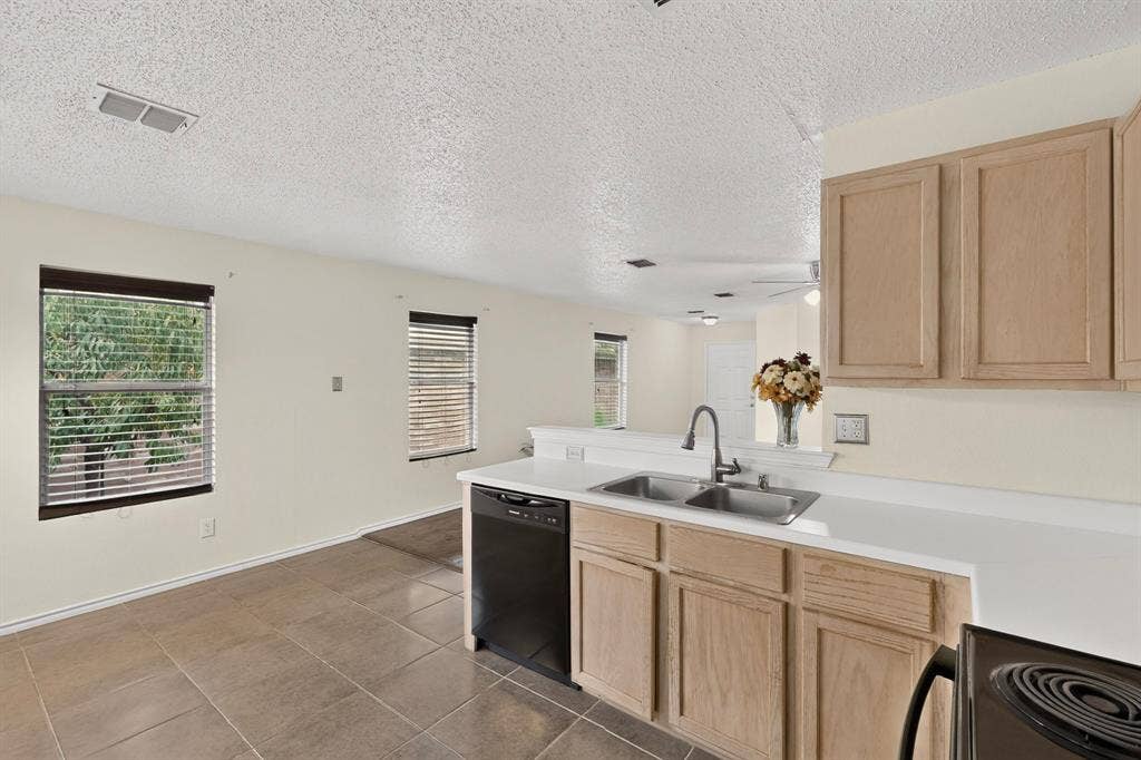 3107 Westview Drive, McKinney, TX 75070 Kitchen featuring light wood-finish cabinetry, solid surface countertops, dual-basin stainless steel sink, black built-in dishwasher, and ceramic tile flooring