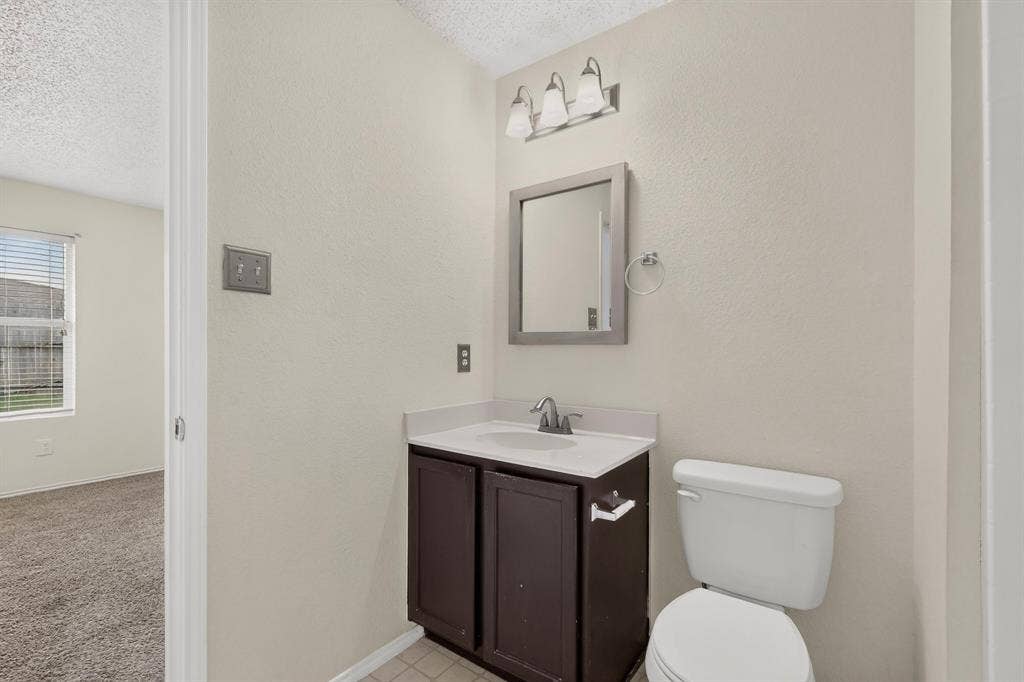 3107 Westview Drive, McKinney, TX 75070 Vanity with dark cabinetry and white countertop, integrated sink, wall-mounted mirror, three-light fixture, and porcelain toilet
